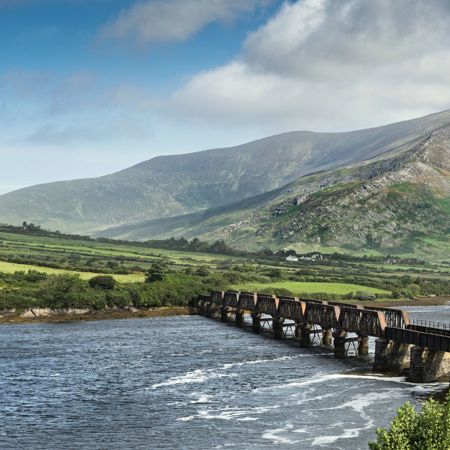 A view of the Caragh River flowing through a valley in County Kerry, Ireland, with mountains rising in the background under a partly cloudy sky