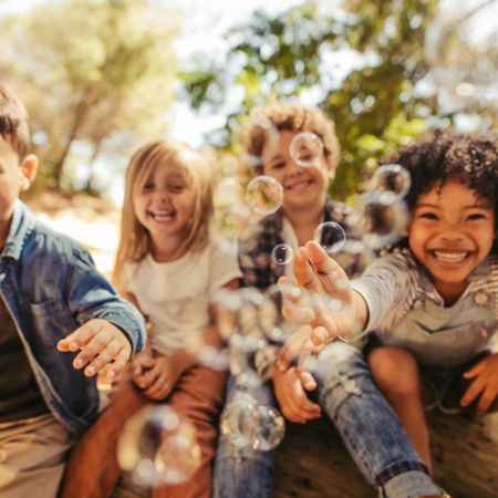 Four children are sitting outdoors, laughing and reaching for bubbles floating in the air