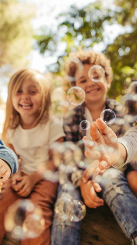 Four children are sitting outdoors, laughing and reaching for bubbles floating in the air